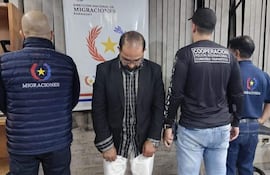 Hombre con chaqueta negra en el centro, rodeado de dos autoridades que llevan chalecos de Migraciones y Cooperación Policial Internacional.