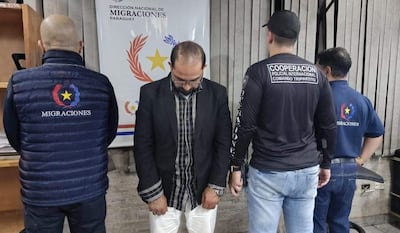 Hombre con chaqueta negra en el centro, rodeado de dos autoridades que llevan chalecos de Migraciones y Cooperación Policial Internacional.