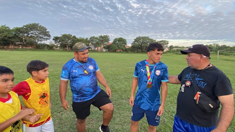 Cuatro personas en un campo de fútbol: un joven con medalla y tres adultos, dos con camisetas azules y negro, conversando en un ambiente familiar.
