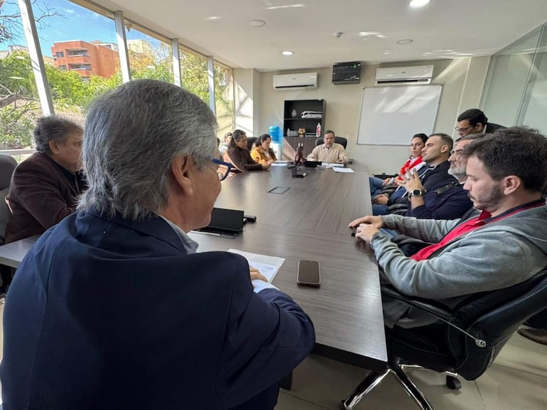 Hombres en reunión con documentos y teléfonos móviles, algunos visten camisetas rojas y negras, ambiente iluminado con plantas.
