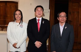 Tres personas conversan: mujer con chaqueta blanca, hombre en traje negro con corbata roja, y otro en traje gris, en ambiente elegante.