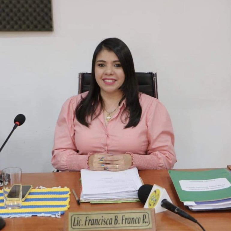 Lic. Francisca B. Franco R. sonríe con blusa rosa en su oficina, rodeada de documentos y objetos de trabajo.