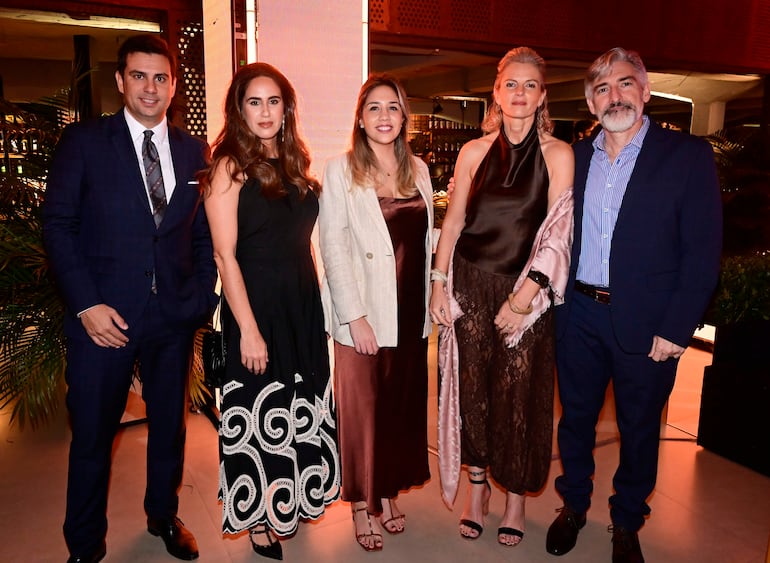 Tres mujeres en vestidos elegantes y dos hombres en traje sonríen en un ambiente festivo con luces decorativas.