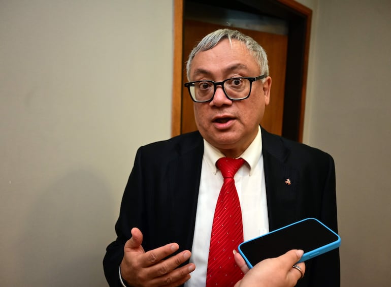 Hombre de cabello gris en abrigo negro y corbata roja, gesticulando mientras habla; mano con teléfono móvil a su lado en un ambiente interior.