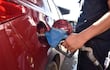 Hombre con camisa oscura y tatuaje en brazo derecho inserta una manguera de combustible en un coche rojo en gasolinera.