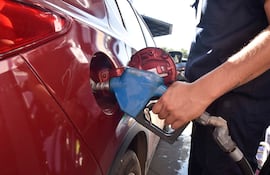 Hombre con camisa oscura y tatuaje en brazo derecho inserta una manguera de combustible en un coche rojo en gasolinera.