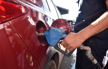 Hombre con camisa oscura y tatuaje en brazo derecho inserta una manguera de combustible en un coche rojo en gasolinera.