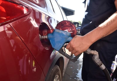 Hombre con camisa oscura y tatuaje en brazo derecho inserta una manguera de combustible en un coche rojo en gasolinera.