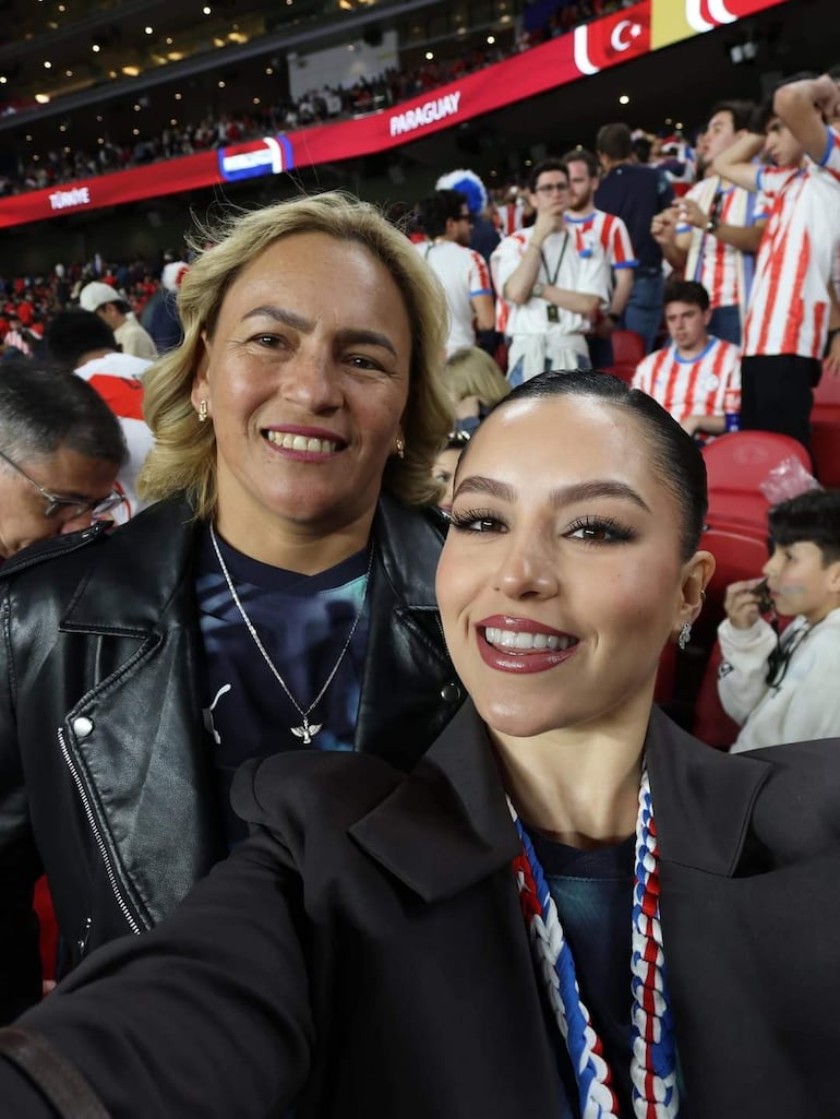 Dos mujeres sonríen, una con chaqueta de cuero y otra con blusa negra, rodeadas de aficionados en un estadio lleno de camisetas rojas y blancas.