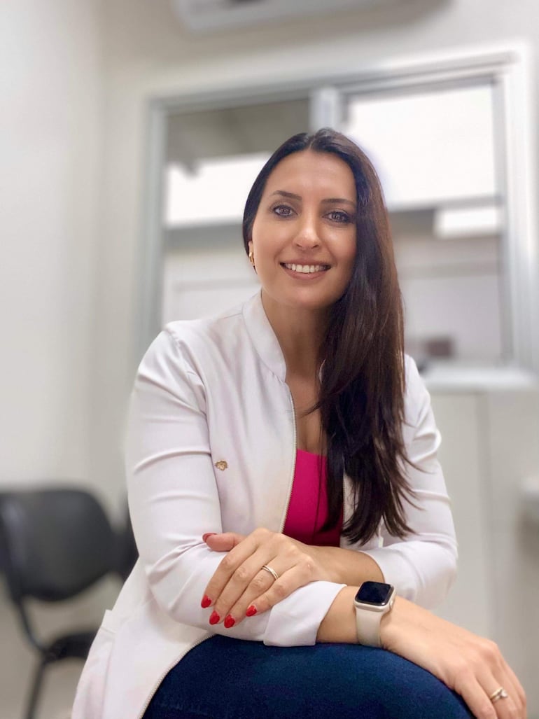 Laura Zalazar luciendo chaqueta blanca y blusa fucsia, sonriendo mientras está sentada en un consultorio médico.