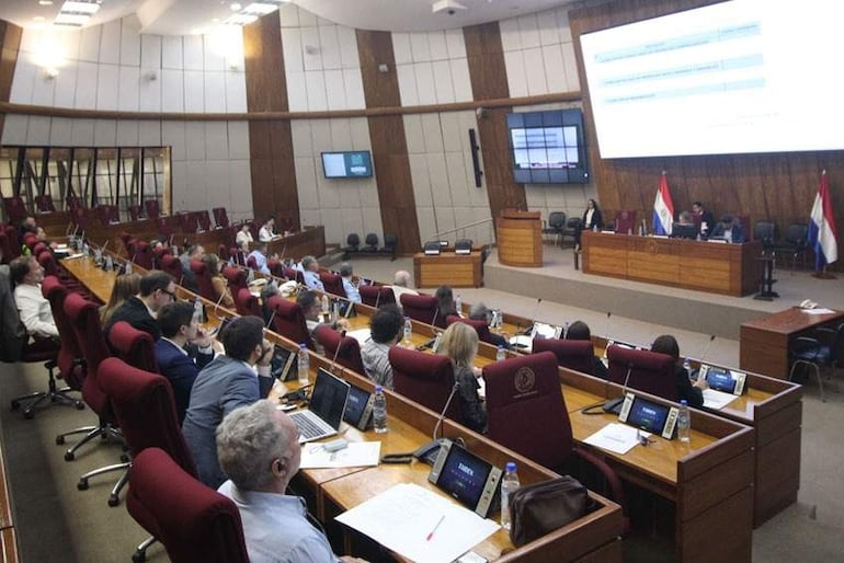 Asistentes en un recinto legislativo con bancos rojos, algunos tomando notas, y pantalla grande mostrando datos relevantes.