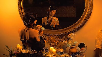 Mujer de espaldas con pelo largo y oscuro, sosteniendo flores en un espejo dorado en un ambiente cálido con velas.