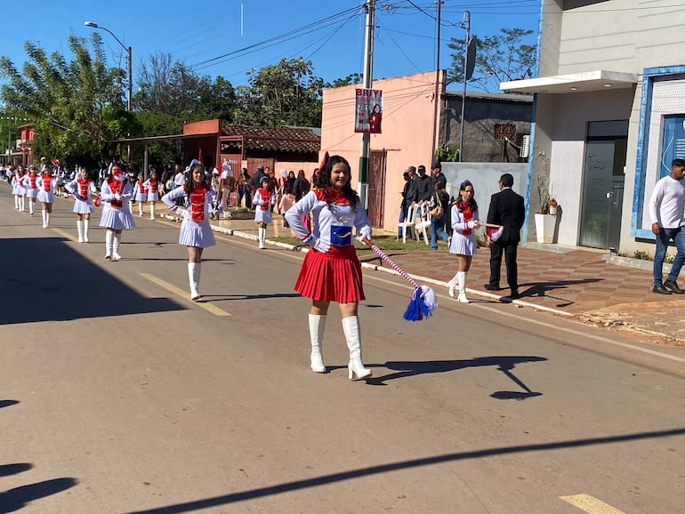 Grupo de jóvenes bailarinas en trajes blancos y rojos desfilan, mientras espectadores observan desde sillas en un ambiente festivo.