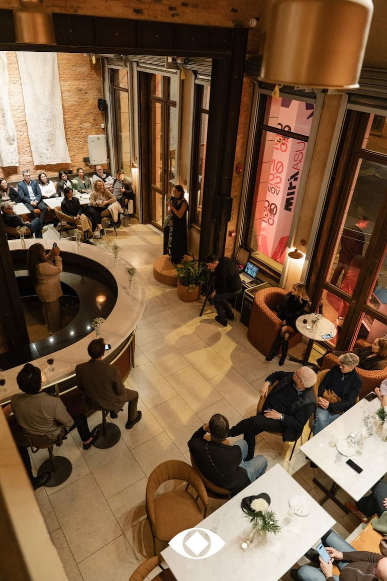 Mujer de pie presentando en un restaurante, asistentes sentados en mesas con atención, decorado contemporáneo y acogedor.
