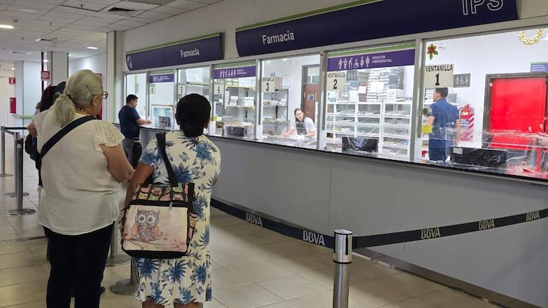 Mujeres en la fila de la farmacia. Una usa lentes y la otra lleva un vestido floral y mochila con diseño de búhos.