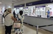 Mujeres en la fila de la farmacia. Una usa lentes y la otra lleva un vestido floral y mochila con diseño de búhos.