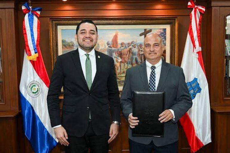 Luis Bello en traje oscuro y corbata verde sonríe junto a un hombre en traje gris claro que sostiene un documento en una carpeta negra.