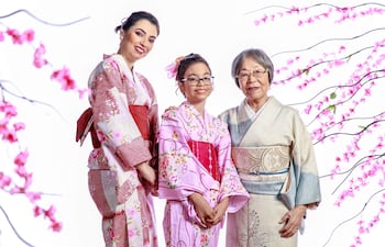 Mujer con kimono rojo, niña en kimono rosa y mujer mayor en kimono gris claro, sonriendo en un entorno festivo con flores de cerezo.