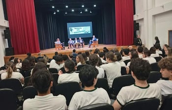 Jóvenes en camisetas blancas con detalles verdes escuchan a oradores en un auditorio con cortinas rojas y proyector iluminado.