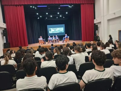 Jóvenes en camisetas blancas con detalles verdes escuchan a oradores en un auditorio con cortinas rojas y proyector iluminado.