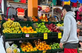 Hombre en sudadera gris y shorts rojos selecciona frutas entre cestas coloridas en un mercado iluminado.