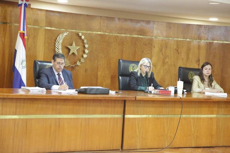 Hombre con traje oscuro escribiendo, mujer rubia hablando con micrófono y otra mujer observando, en un entorno formal con bandera paraguaya.