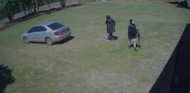 Dos personas con chaquetas oscuras en un jardín, una sostiene un objeto que parece un arma, ambiente tranquilo con césped y vehículo plateado.