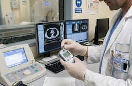 Médico en uniforme blanco y guantes, sosteniendo un dispositivo en un laboratorio con monitores que muestran imágenes médicas.