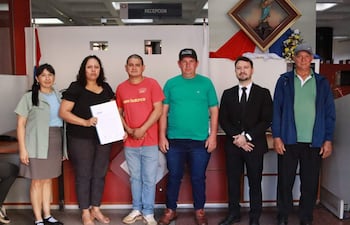 Mujer en blusa negra sostiene un documento mientras hombres la observan en oficina con mobiliario visible.