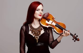 Rachel Kolly con cabello largo y rojo en un elegante vestido negro, sosteniendo un violín contra un fondo gris.