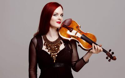 Rachel Kolly con cabello largo y rojo en un elegante vestido negro, sosteniendo un violín contra un fondo gris.