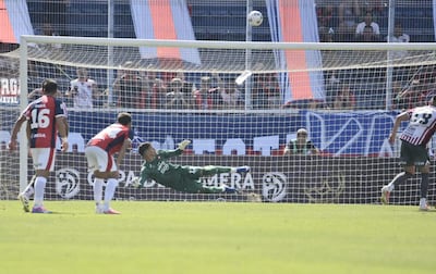 Portero de uniforme verde en acción, en un partido de fútbol, con público y una gran bandera azul y roja de fondo.