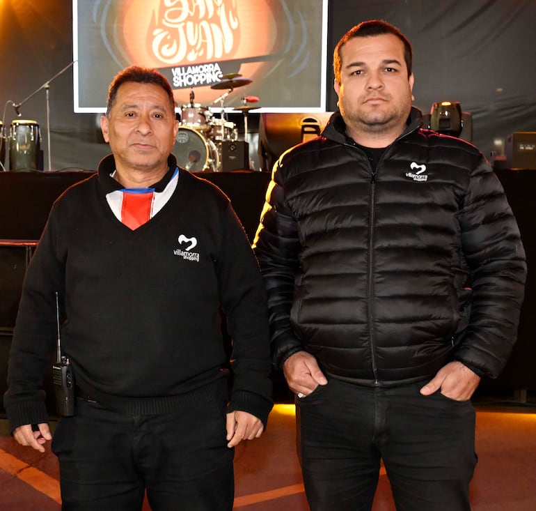 Dos hombres, uno con suéter negro y otro con chaqueta acolchada, observan un escenario con equipo musical.