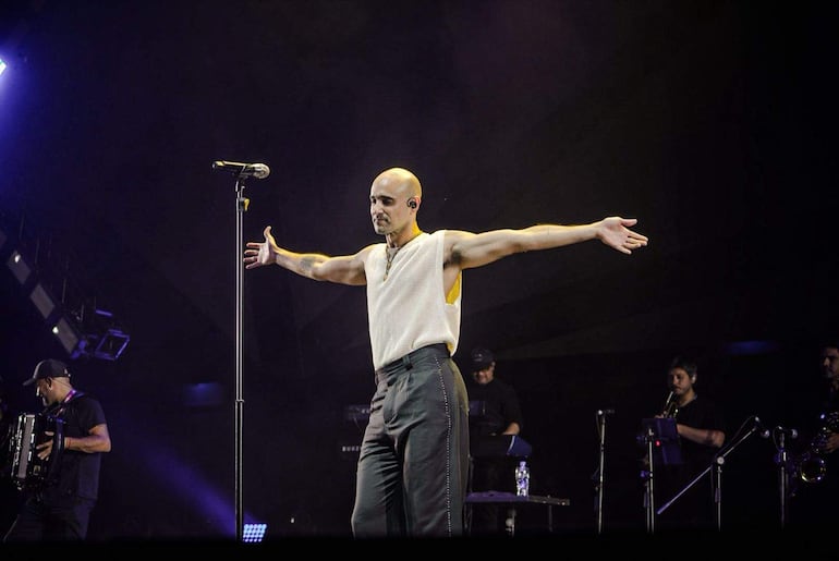 Abel Pintos, con camiseta de malla blanca, interactúa con el público mientras músicos lo acompañan en el escenario iluminado.
