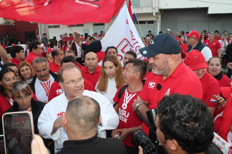 Camilo Pérez habla enérgicamente rodeado de hombres con camisetas rojas y banderas en un evento político al aire libre.