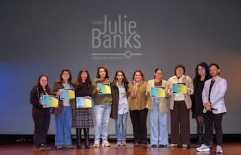 Ocho personas sonrientes en un escenario sosteniendo diplomas coloridos, con un hombre a la derecha hablando al público.