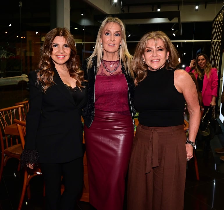 Tres mujeres posan sonrientes en un salón iluminado, luciendo vestimentas elegantes de colores variados.