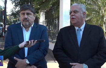 Dos hombres en trajes formales, uno azul y otro negro, discuten mientras uno sostiene un teléfono móvil.