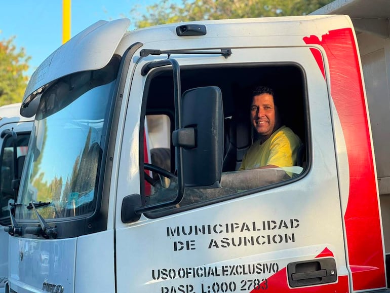 Hombre sonriendo al volante de un camión municipal de Asunción, vestido con camiseta amarilla y vegetación de fondo.