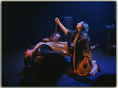 Tres mujeres en escena teatral: una acostada en la mesa, otra sosteniéndola y una tercera de pie, todas con expresiones dramáticas.