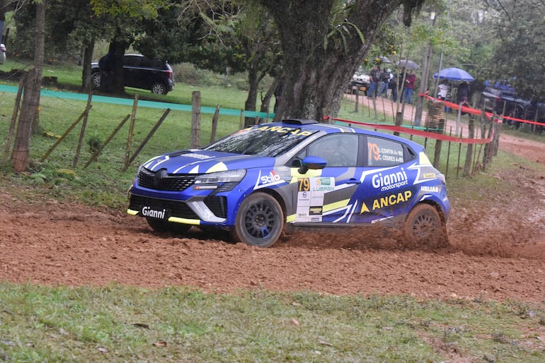 Auto Renault decorado en azul y amarillo de Gianni Motorsport, levantando tierra en el rally, con público bajo paraguas al fondo.