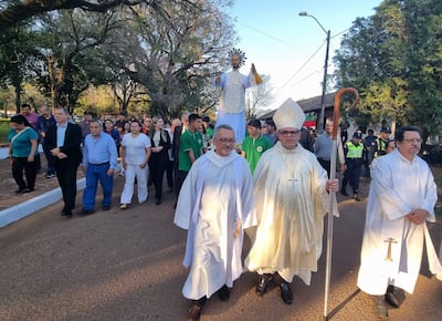 Dos sacerdotes en vestimenta blanca y dorada lideran una multitud en procesión, mientras otros asistentes con ropa formal los acompañan.