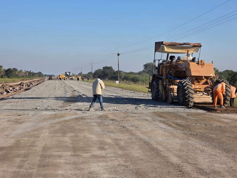 Dos trabajadores en abrigo claro y jeans, uno usa una herramienta y otro opera una motoniveladora en un entorno de construcción.