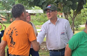 Elías Cabral viste camisa blanca y gorra colorida, mientras saluda a un hombre camiseta naranja. Ambiente de evento comunitario.