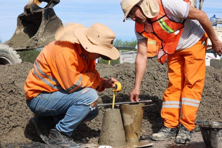 Dos trabajadores en chalecos naranjas y sombreros, uno midiendo con cinta métrica, el otro alisando concreto en obra al aire libre.