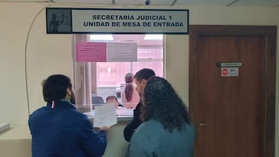 Hombre de espaldas con chaqueta azul sostiene documentos, mientras otra persona interactúa con el ventanuco en oficina judicial.