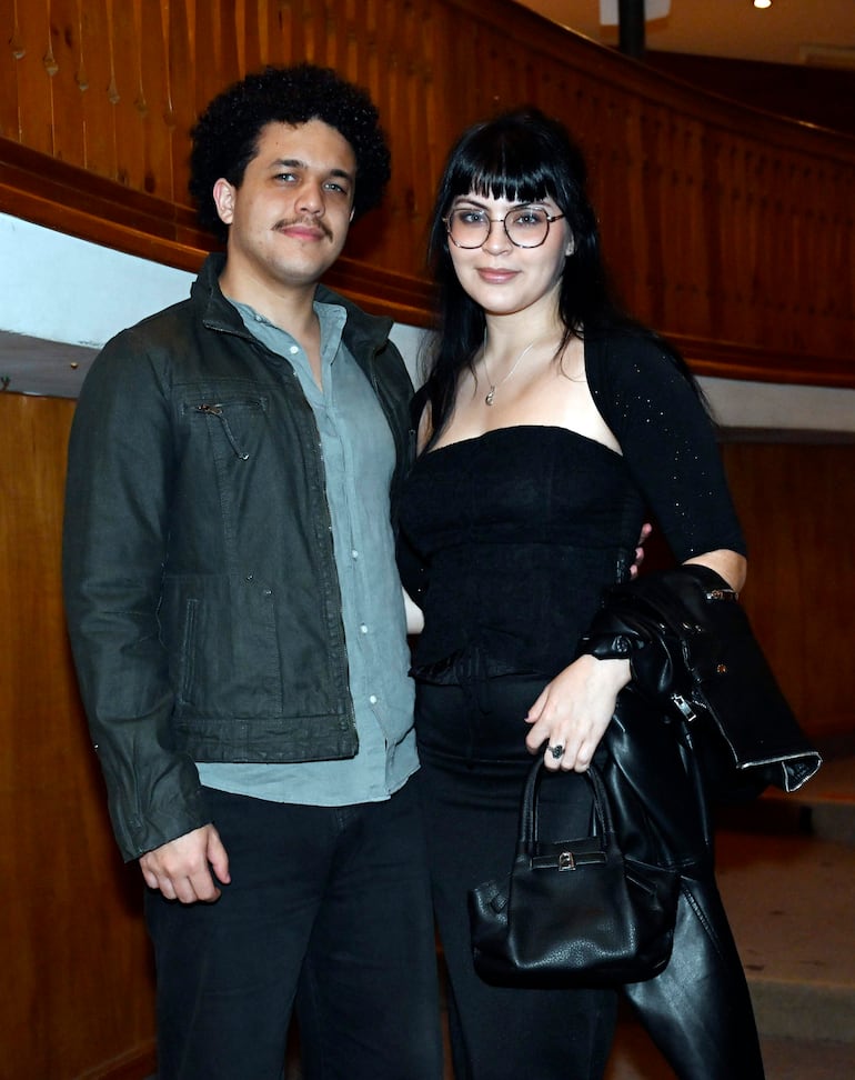 Hombre con chaqueta oscura y mujer con vestido negro posan juntos en un elegante ambiente teatral con estructura de madera al fondo.