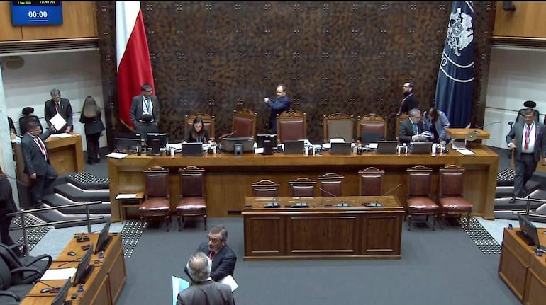 Mujer en computadora portátil y hombres de pie en el Senado de Chile, con iluminación cálida y banderas al fondo.