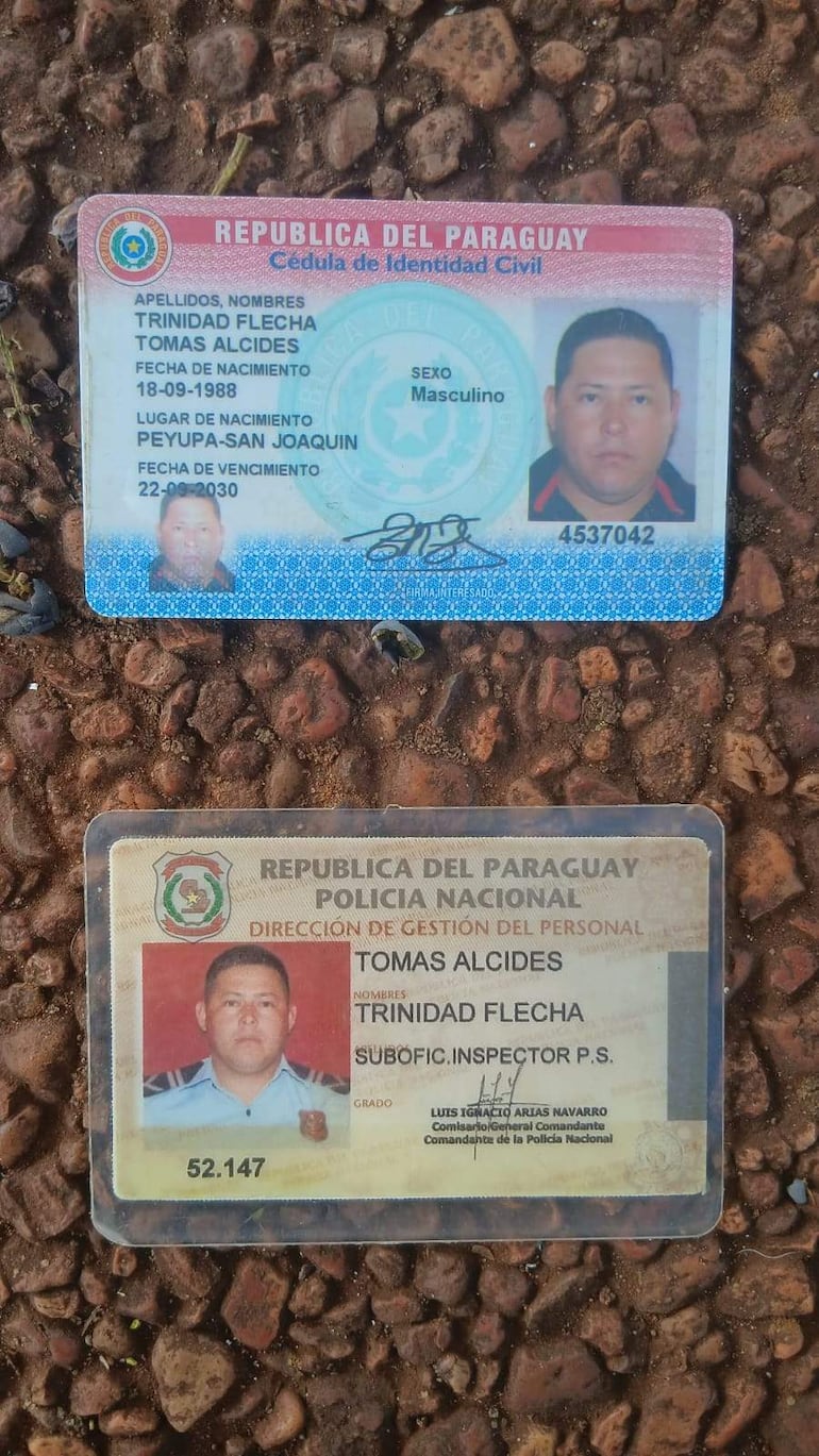 Cédulas de identidad civil y policial de Trinidad Flecha sobre fondo de tierra y guijarros, con detalles visibles de los documentos.