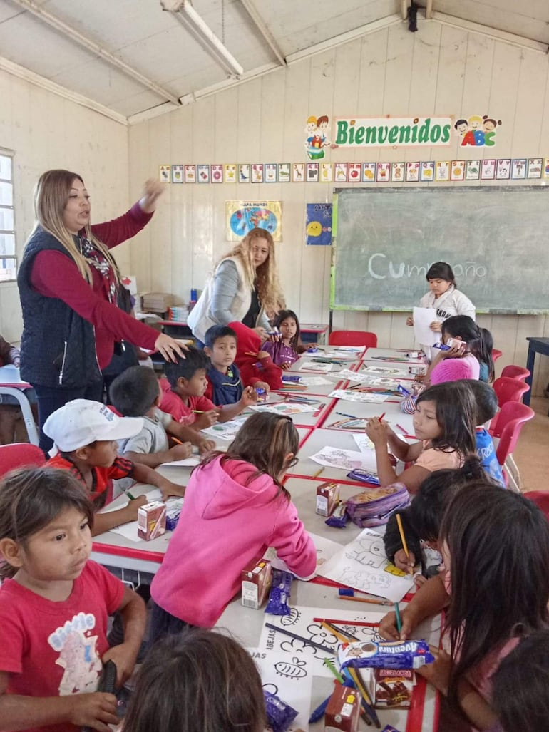 Niña con camiseta de colores dibuja en mesa, adultos interactúan con los estudiantes en aula con letrero de bienvenida.